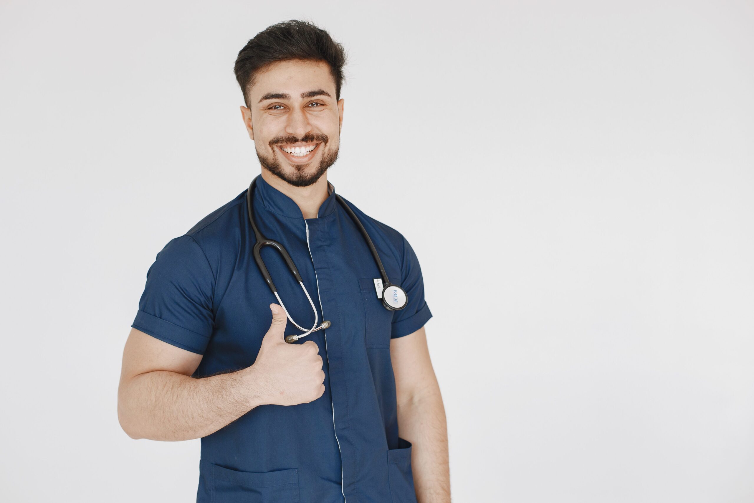 À propos Confident male doctor in blue scrubs with a stethoscope smiling and giving a thumbs up.